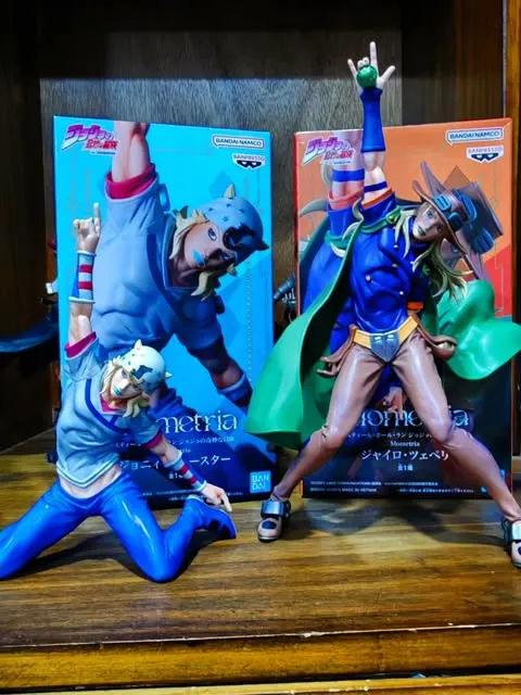 Las figuras de Johnny Joestar y Gyro Zeppeli fuera de sus cajas, sobre una repisa de madera.